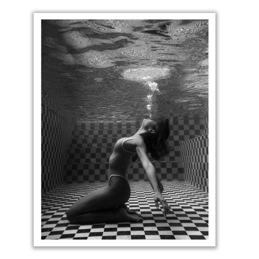 A woman wearing a swimsuit kneels on the checkered floor at the bottom of a swimming pool, arching her back and exhaling bubbles towards the water's surface. This black and white image, titled "The Banya #2" by Francesca Owen, is available on fine art paper, comes hand signed with worldwide shipping options.