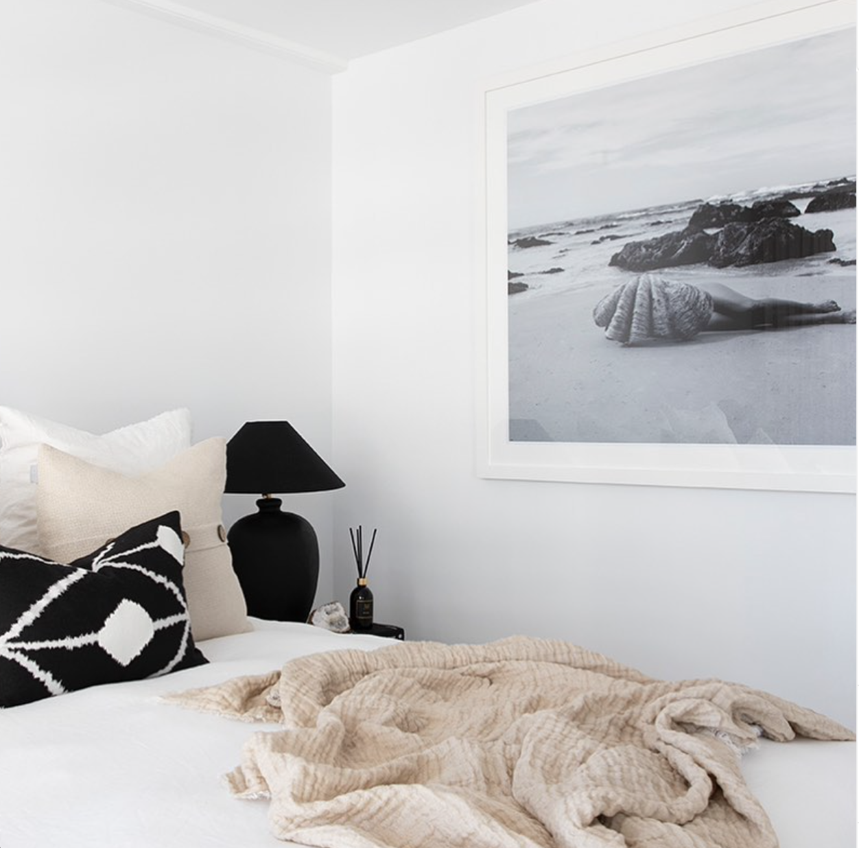 A modern bedroom showcases a neatly made bed adorned with a beige textured blanket, white pillows, and a black-and-white geometric cushion. On the nightstand beside the bed sits a sleek black table lamp. Above, an elegant fine art photograph titled "The Beauty Within B&W" by Francesca Owen of a snail on a beach adds sophistication to the space.