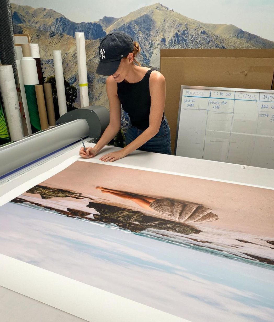 A person in a black tank top and blue jeans, wearing a black cap, is working with "The Beauty Within" by Francesca Owen on a flatbed printer using archival pigments. The image features a pink sand beach and hills. Rolled prints and a whiteboard are visible in the background.