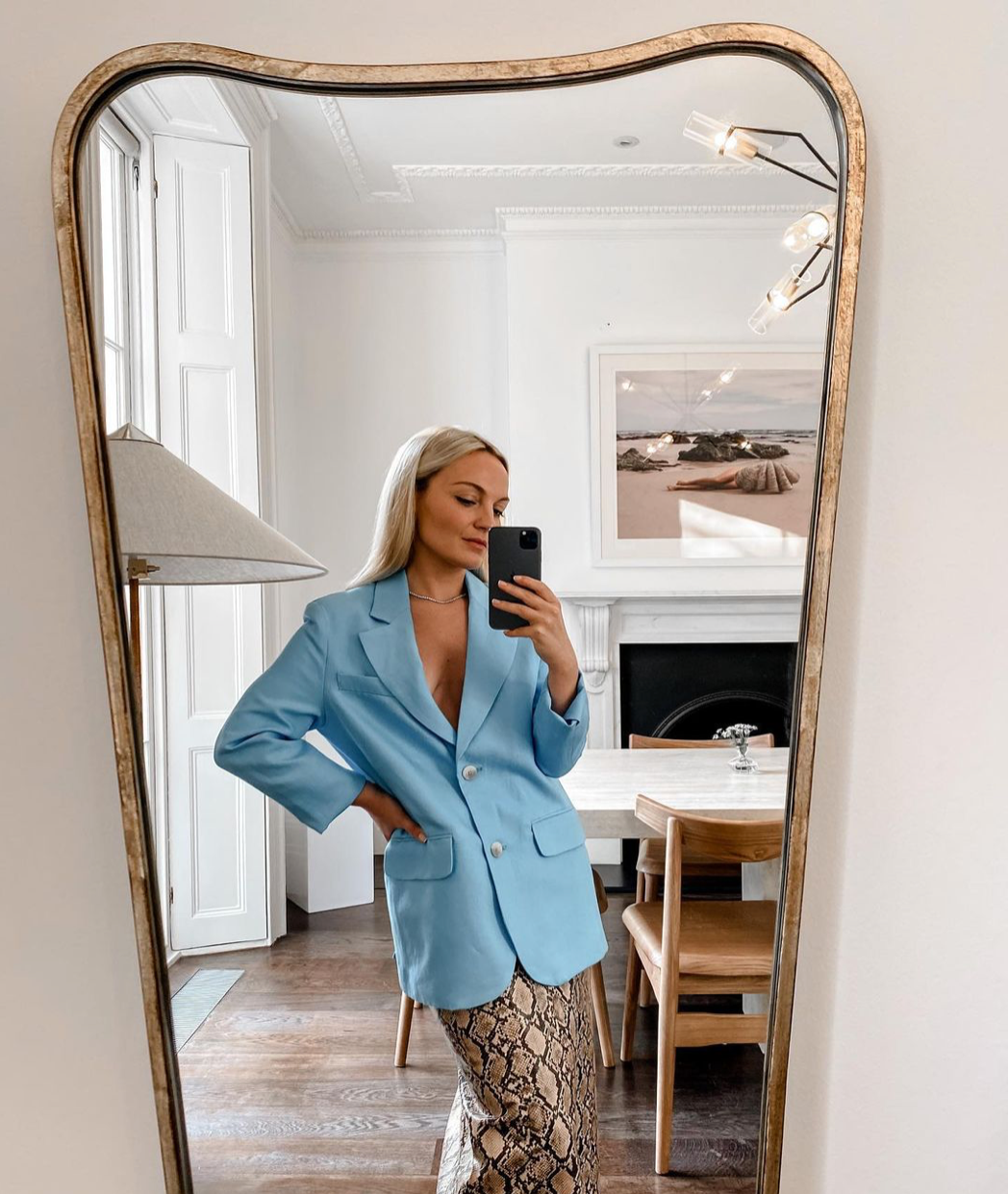 A person with long, light hair takes a selfie in a large, ornate mirror. They are wearing a light blue blazer over a patterned skirt. The room behind features a dining table with chairs, a floor lamp, white walls, and "The Beauty Within," an artwork by Francesca Owen, printed on fine art paper using archival pigments and elegantly positioned above the fireplace.