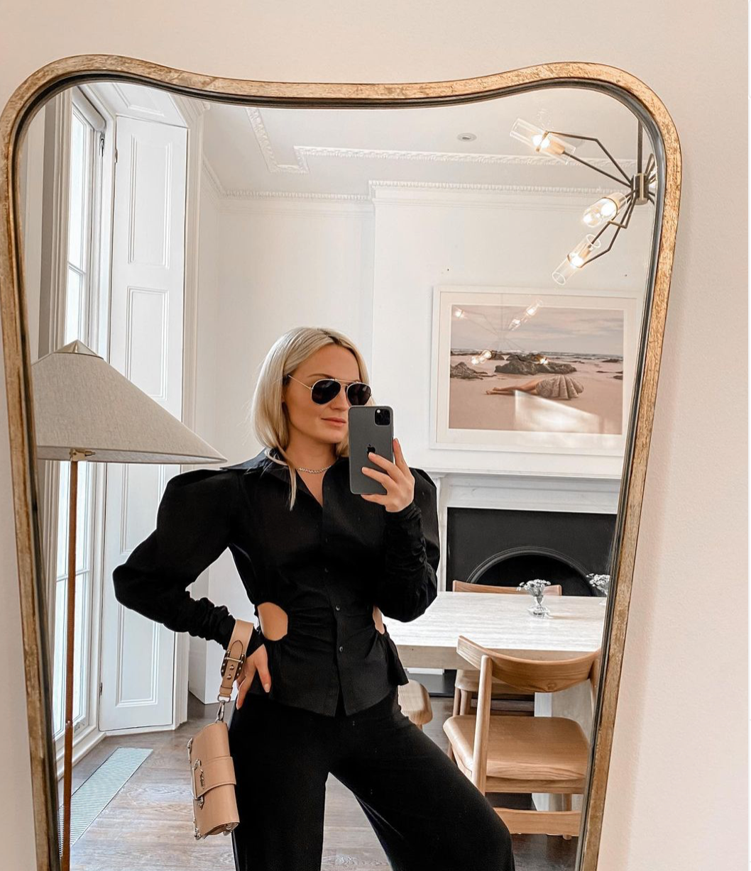 A person with blonde hair and sunglasses takes a mirror selfie in a stylish room. They are wearing "The Beauty Within," a black top with cut-out sections at the sides, designed by Francesca Owen, and holding a beige handbag. The room features a wooden table with chairs, a tall white lamp, and framed fine art paper photographs on the wall.