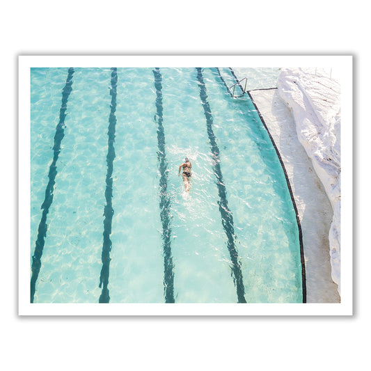 A person is swimming in a large, clear Bondi outdoor pool by Francesca Owen, heading towards the far end. The pool is divided into several swimming lanes by dark lines on the pool floor. The sunlight casts a ripple effect across the water, resembling an intricate pattern on fine art paper. White Bondi pool tiles are visible on the right side.