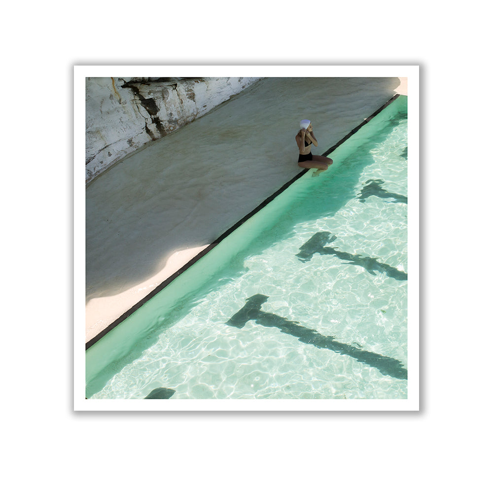 A person wearing a black swimsuit and white hat sits on the edge of a clear turquoise swimming pool, their feet dipping into the water. A rocky, sunlit wall serves as a striking backdrop. The scene evokes the timeless elegance of fine art prints, reminiscent of Francesca Owen's "Bondi Empty Lanes," with shadows cast by the pool's metal steps creating an almost archival pigments masterpiece.