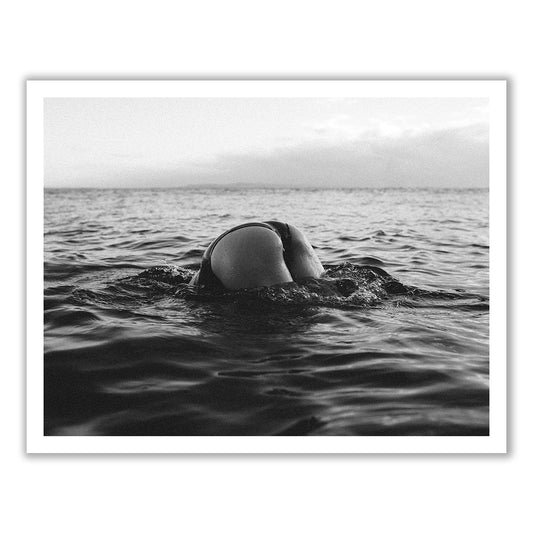 Black and white photo titled "Bottoms Up" by Francesca Owen, showcasing a person diving underwater in the ocean with only their lower body in swimwear visible above the surface, against a horizon backdrop. This stunning image is available as fine art prints, crafted with archival pigments to ensure lasting quality.