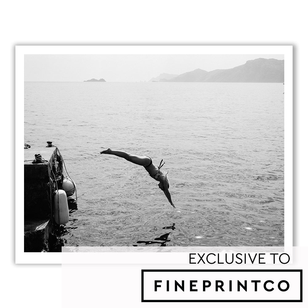 Dive In" by Francesca Owen showcases a person diving into a serene body of water from a dock, where a small boat is moored. The soft outlines of distant islands or landmasses can be seen in the background. This fine art print is created using archival pigments on fine art paper and includes a certificate of authenticity.