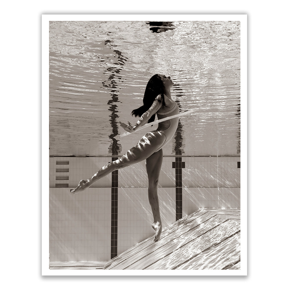 Flying Solo 02 by Francesca Owen captures a graceful underwater photograph in black and white of a woman in a one-piece swimsuit, performing a ballet-like pose with one leg extended and arms outstretched, immersed in a swimming pool. Light ripples and reflections create a serene and artistic effect, expertly printed on fine art paper with archival pigments.