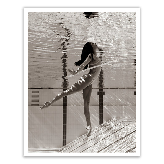 Flying Solo 02 by Francesca Owen captures a graceful underwater photograph in black and white of a woman in a one-piece swimsuit, performing a ballet-like pose with one leg extended and arms outstretched, immersed in a swimming pool. Light ripples and reflections create a serene and artistic effect, expertly printed on fine art paper with archival pigments.