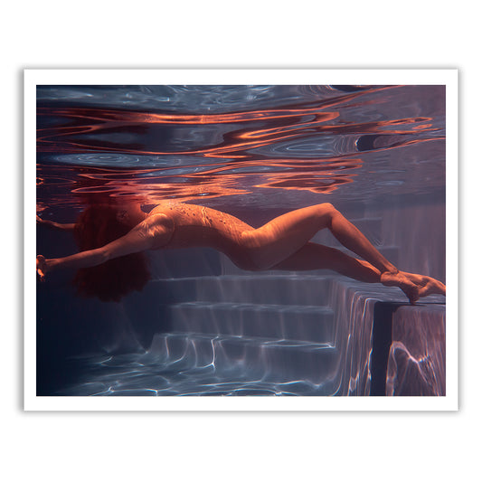 A person with curly hair floats effortlessly underwater in a pool, wearing a one-piece swimsuit near the steps. Light patterns dance on the water and reflect off the pool walls, creating a dreamlike scene perfect for fine art prints. Each detail is captured with archival pigments under the brand Francesca Owen's Glisten 08 collection, and custom framing ensures timeless beauty.