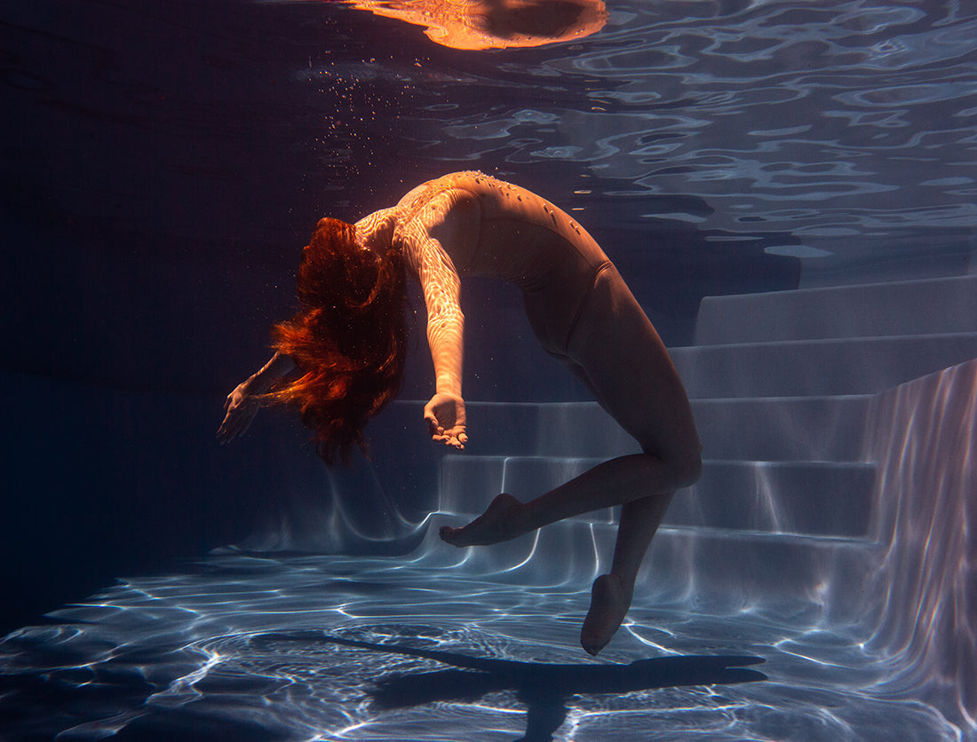 A woman with long hair floats gracefully underwater, arching her back with arms extended, adorned in a form-fitting outfit from Francesca Owen's Glisten 02 series. She is immersed in a serene pool where gentle light patterns dance on the water, and steps are visible in the background. This stunning image is available as fine art prints with worldwide shipping.