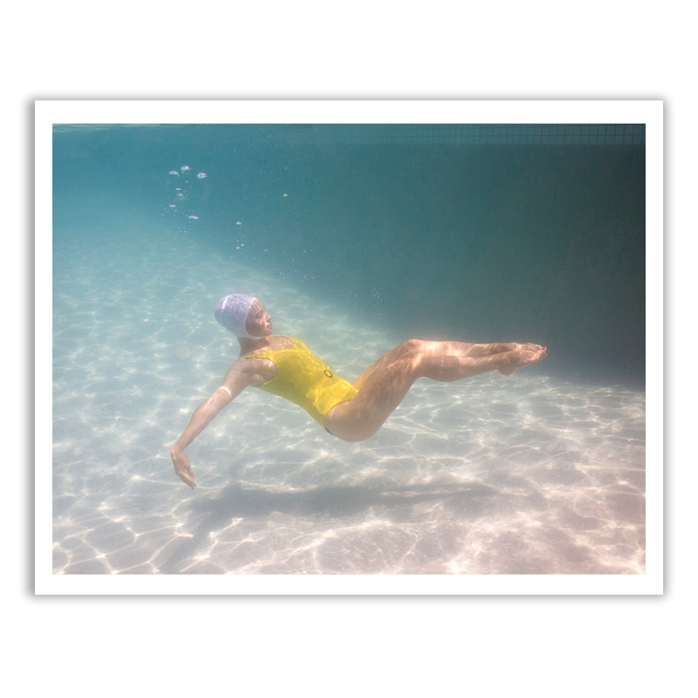 A person wearing the Francesca Owen Halcyon Daze 2 yellow swimsuit floats underwater in a pool, with sunlight casting patterns on the pool floor. The person’s arms are extended gracefully as small air bubbles ascend towards the surface. The clear blue water captures this serene moment, perfect for fine art prints.