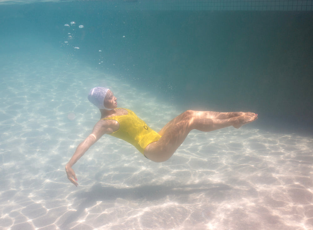 A person in a yellow swimsuit and white swim cap is floating underwater in a swimming pool. The sun's rays create ripples of light on the pool floor, and the water appears calm and clear. The scene, reminiscent of a Francesca Owen fine art print titled "Halcyon Daze 2," captures the individual gracefully extending their arms in a horizontal position.