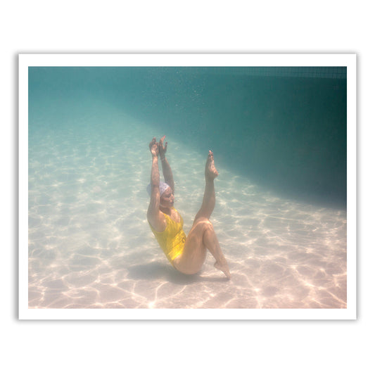 A person wearing the Halcyon Daze 3 yellow swimsuit by Francesca Owen performs a synchronized swimming move underwater, with legs stretched upward and arms bent at the elbows. The sunlight creates dappled patterns on the pool floor, resembling the delicate touch of archival pigments used in fine art prints.