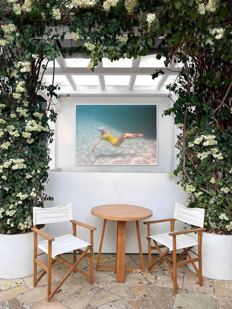 A small round wooden table with two white chairs sits under a pergola with flowering vines. On the wall is "Halcyon Daze #2," a hand-signed art print by Francesca Owen, depicting a swimmer underwater in a yellow swimsuit.