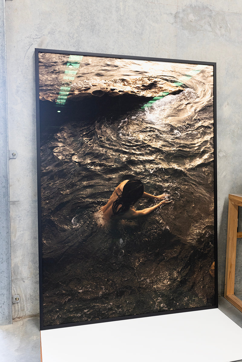 A framed photograph titled "Sunrise Swim #9" by Francesca Owen, leaning against a wall, depicts a person with long hair swimming in a body of water surrounded by rocky terrain. The water appears to be in motion, creating ripples and waves around the swimmer. Printed with archival pigments, it includes a certificate of authenticity.