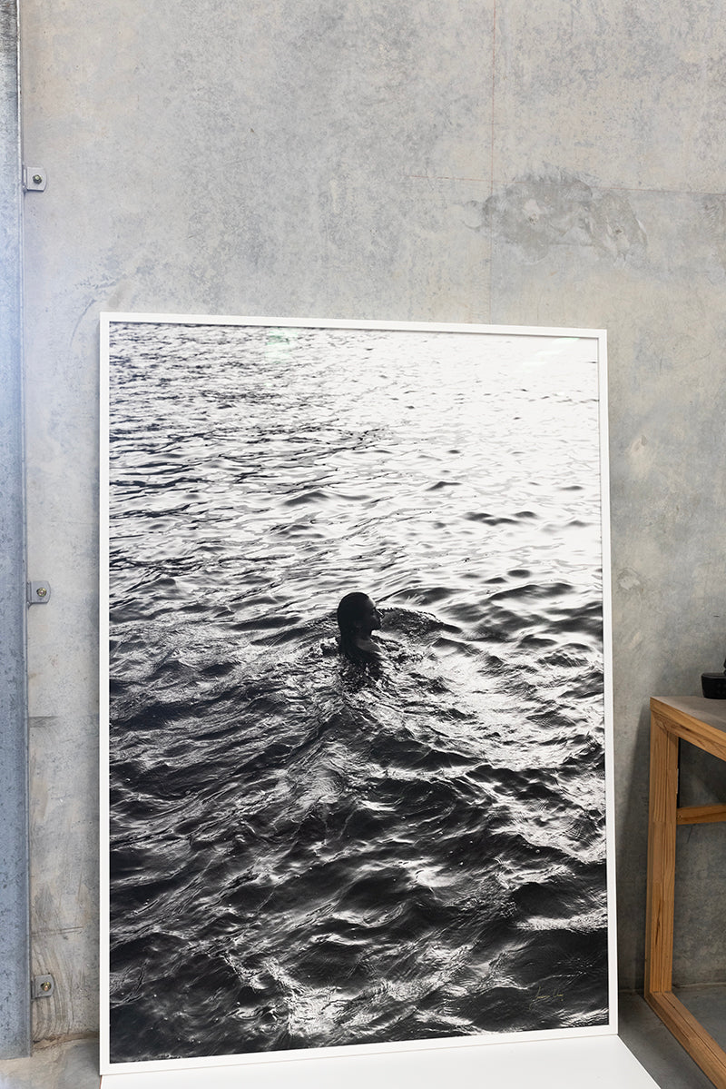 A black-and-white photograph from the "Sunrise Swim #3" collection by Francesca Owen leans against a concrete wall, depicting a solitary person swimming in rippled waves that reflect light. Printed using archival pigments, this fine art print captures timeless beauty. A wooden table with a book is partially visible on the right.