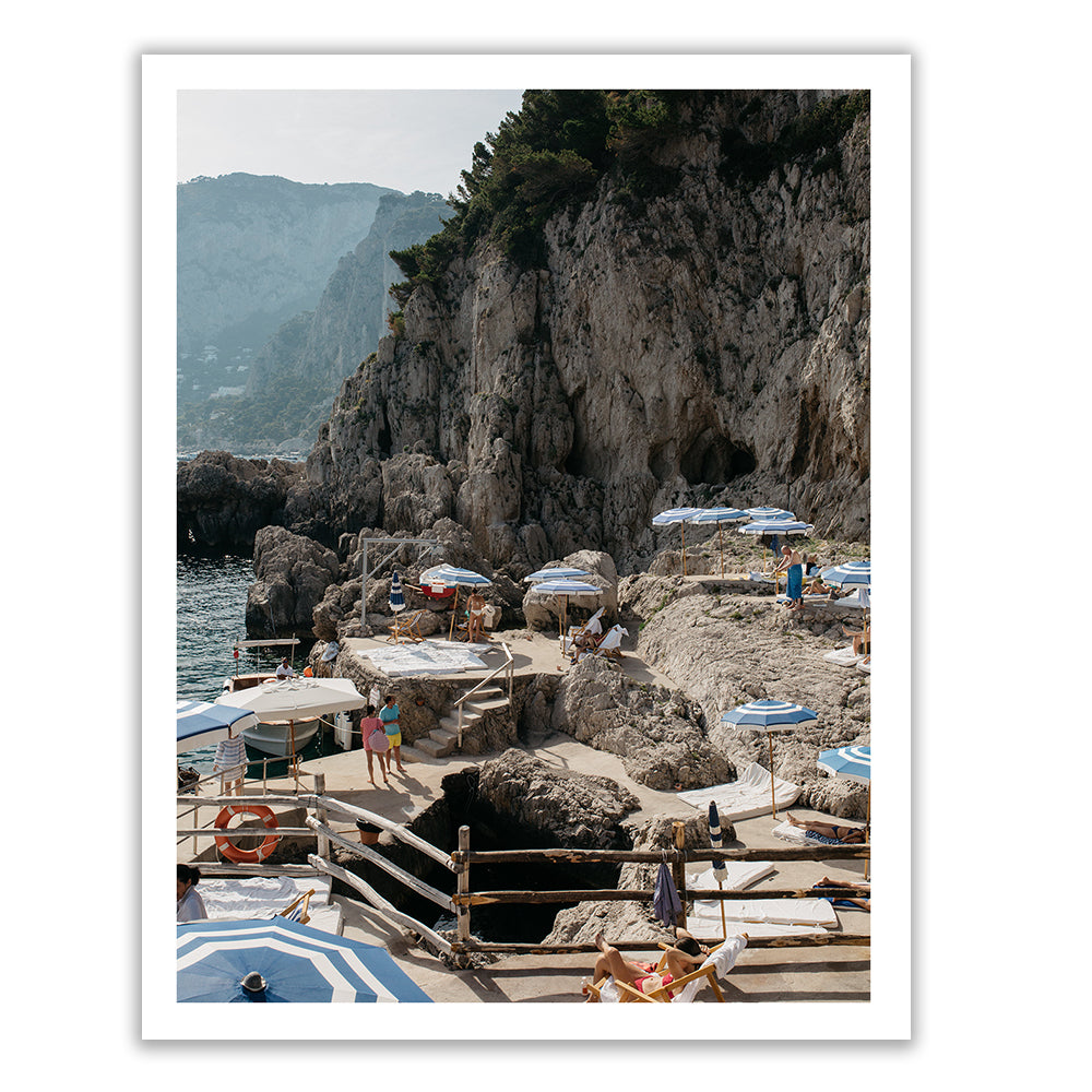 Scenic coastal view of La Fontelina #1 by Francesca Owen, depicting a rocky beach with several people under blue and white umbrellas. Steep cliffs and green trees loom in the background. The calm sea and rocky shoreline, available in custom sizes on fine art paper, feature various sunbathing areas and a few swimmers.