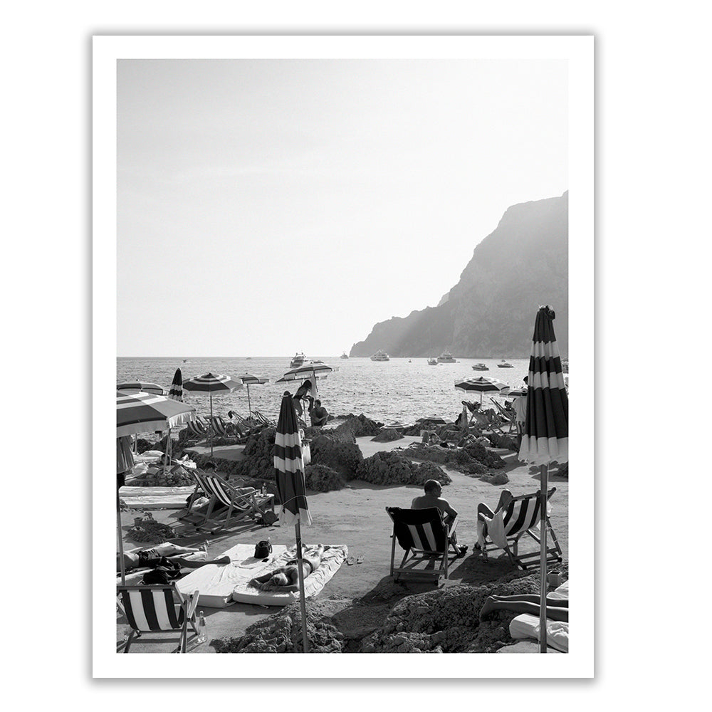 La Fontelina #2 by Francesca Owen is a stunning black and white photograph of a rocky beach scene. People relax on sun loungers under umbrellas, captured with archival pigments on fine art paper. Boats are visible on the water, while jagged cliffs rise from the sea in the background.