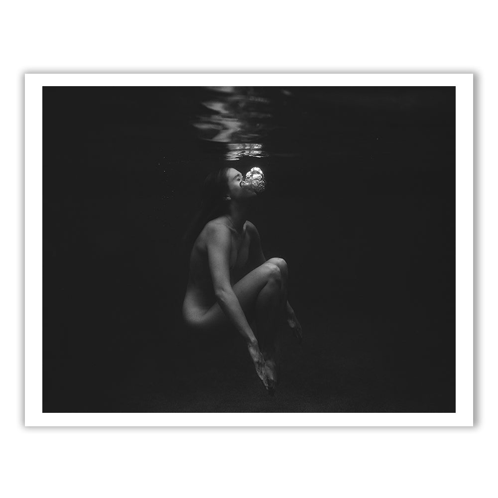 The black and white underwater photograph titled "Let Go" by Francesca Owen captures a person sitting in a fetal position, floating just below the water's surface. Bubbles escape from their mouth, rising towards the surface, while dim lighting casts a serene and reflective mood. This piece is available on fine art paper with archival pigments in custom sizes.