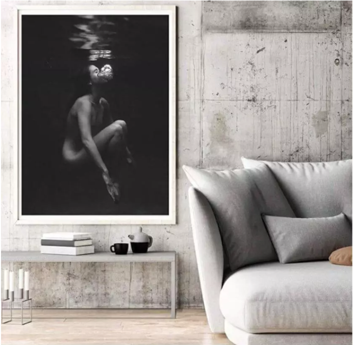A modern living room featuring Francesca Owen's fine art print "Let Go," depicting a person partially submerged in water. The room has a concrete wall, a light gray sofa adorned with throw pillows, and a minimalist coffee table topped with stacked books and a small bowl. A candle holder sits by the sofa.