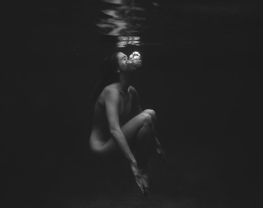 The captivating black and white photograph titled "Let Go" by Francesca Owen depicts a person underwater in a seated position, legs drawn up and hands resting on their knees as they gently blow bubbles that float towards the water's surface. This stunning image is available in custom sizes, printed on Fine Art paper with archival pigments.