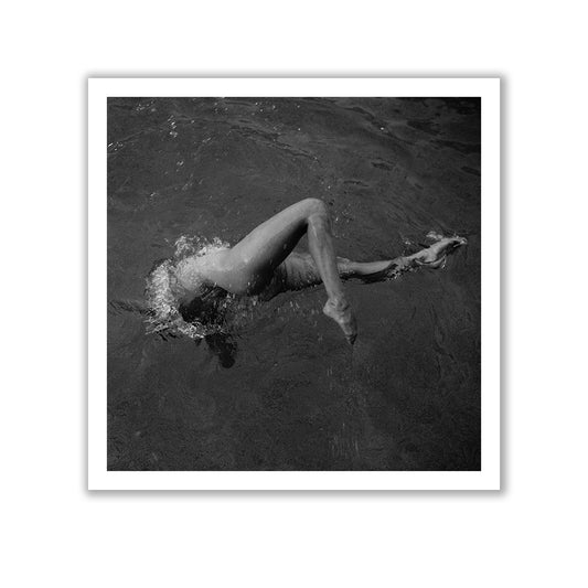 A minimalist interior wall features a framed black-and-white photograph titled "Liquid Legs" by Francesca Owen, depicting a person swimming. Next to the photo, which includes a certificate of authenticity, is a white vase containing sparse green foliage, while a leafy plant sits in the foreground. A dark curtain partially covers a window on the left.