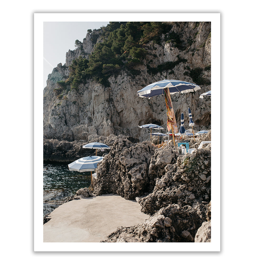 A rocky coastline adorned with blue and white umbrellas shading tables and chairs is framed by majestic, tree-covered cliffs. Sunlight dances across the scene, casting shadows that enhance the tranquil seaside atmosphere. This picturesque view is perfect for immortalization on "Meet Me Here" by Francesca Owen, available on fine art paper or canvas with archival pigments for enduring beauty.