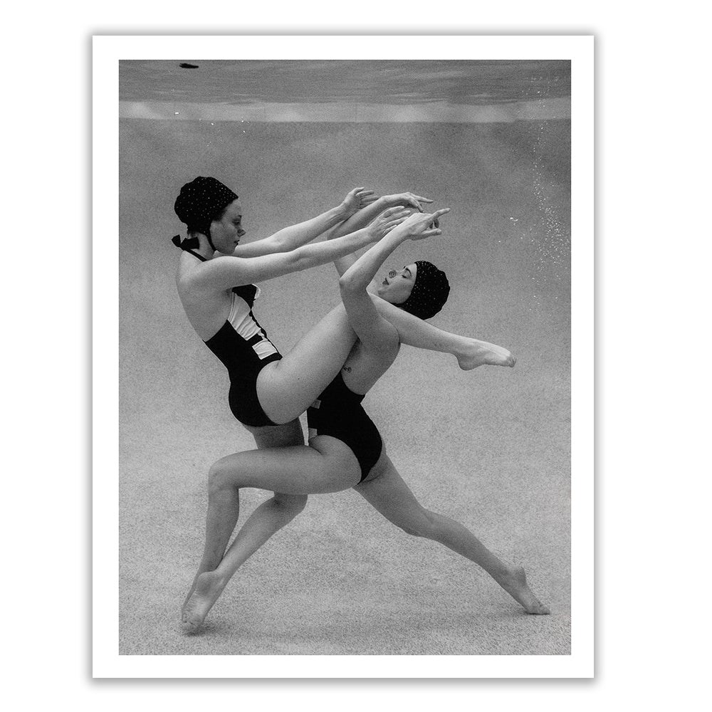 Palm Springs Duo #1 by Francesca Owen is a black and white photo of two synchronized swimmers underwater, clad in swimsuits and swim caps, executing an intricate pose with one swimmer lifting the other. Printed on fine art paper with archival pigments, this image beautifully captures their grace and balance in a suspended, aquatic moment.