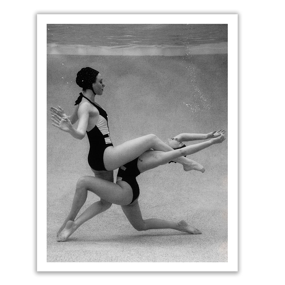 An art display featuring two large, framed black-and-white photographs from the collection "Palm Springs Duo #2" by Francesca Owen, depicting synchronized swimmers underwater. The swimmers wear swim caps and one-piece bathing suits, striking acrobatic poses. The custom-framed photos are mounted on a concrete wall with a shelf below holding books and two cups.