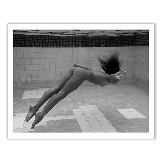 Presenting "Pike" by Francesca Owen: a stunning black and white photograph capturing a person swimming underwater in a tiled pool. The swimmer is streamlined, with arms at their sides and hair flowing upwards, appearing mid-dive and moving gracefully through the water. This piece is available in custom sizes and printed on fine art paper for a timeless finish.