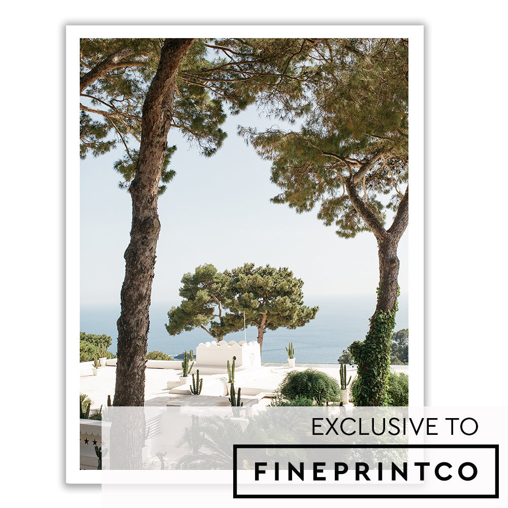 A scenic view featuring tall pine trees framing a white Mediterranean-style building with a backdrop of the blue ocean and distant shoreline. Printed on fine art paper using archival pigments, the "Capri Cacti" by Francesca Owen captures cacti and lush greenery that add to the serene and picturesque atmosphere.