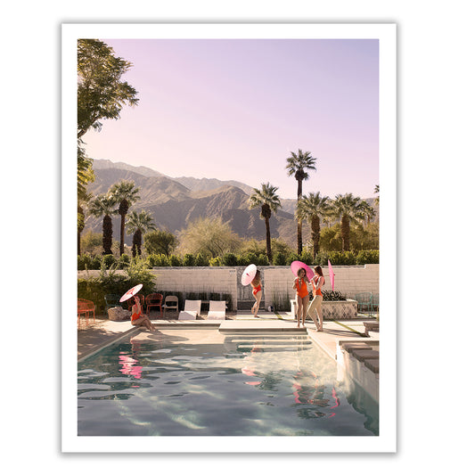 The fine art photograph titled "Poolside Mermaids" by Francesca Owen showcases four women near a poolside, holding parasols. Palm trees and mountains create a scenic backdrop under a pink sky. This limited edition print is elegantly signed in the bottom right corner.