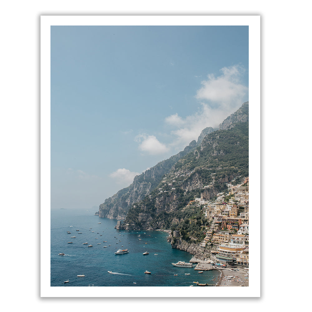 The "Positano #1" by Francesca Owen captures a stunning coastal view of a Mediterranean village perched on steep cliffs, where numerous boats dot the tranquil turquoise waters. The lush green mountains seamlessly blend with a sky adorned with fluffy clouds. This artwork is available in custom sizes to perfectly fit your space.