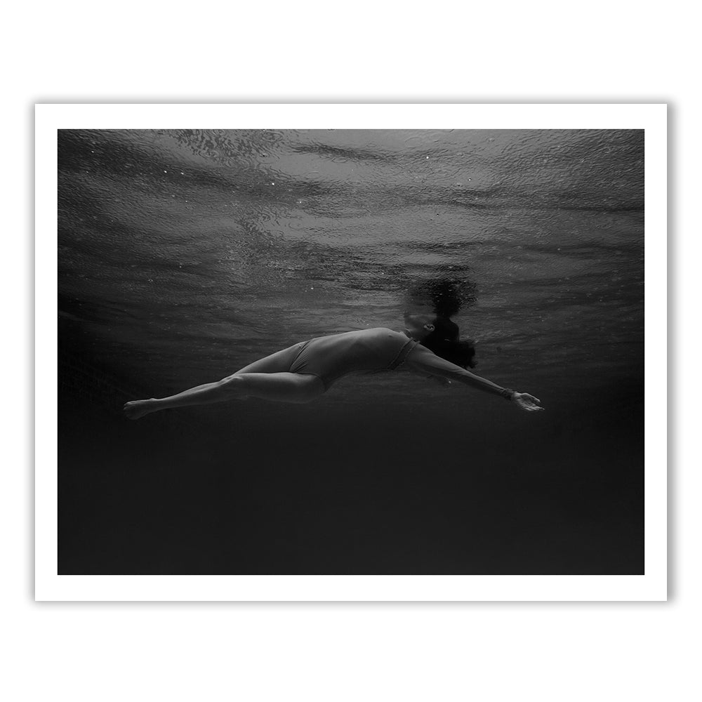 Francesca Owen's Red Head Series #10 features a black and white photograph of a person swimming underwater, stretched out horizontally with arms extended forward and legs straight back. The serene, floating moment is captured on fine art paper and enhanced with archival pigments, contributing to its timeless quality.
