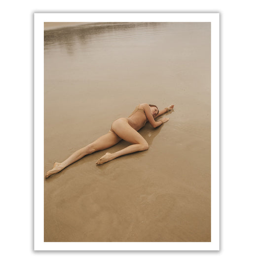 A person from the Francesca Owen's "Red Head Series #3" is lying on a sandy beach near the water's edge. The individual is positioned on their side, with their right arm extended and head resting on it. The scene exudes serenity with neutral, earthy tones prevailing throughout the image, making it an ideal choice for fine art prints using archival pigments.