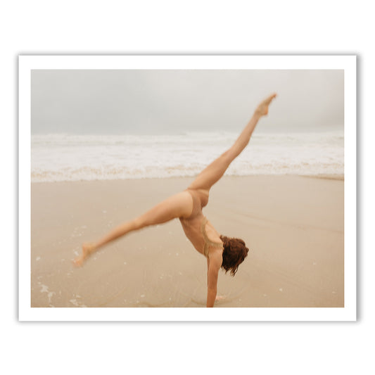 A person in a swimsuit, embodying the essence of Francesca Owen's Red Head Series #4, performs a cartwheel on a sandy beach with waves gently crashing in the background. The overcast sky creates a serene and calm atmosphere, reminiscent of scenes captured on fine art paper with archival pigments.