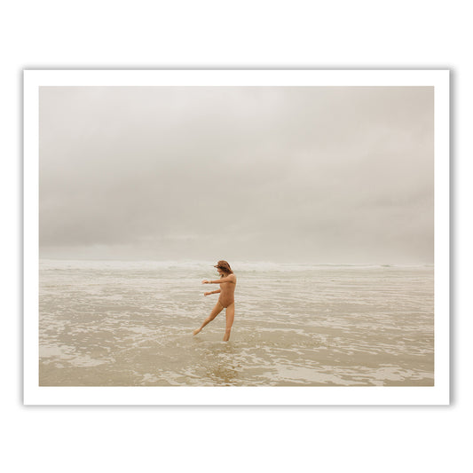 A serene and natural scene is captured in *Red Head Series #5* by Francesca Owen, depicting a nude individual standing in shallow waters on a beach. The person, with arms outstretched, faces slightly to the left amidst gentle waves under a cloudy, overcast sky. This artwork utilizes archival pigments on fine art paper to bring the moment to life.