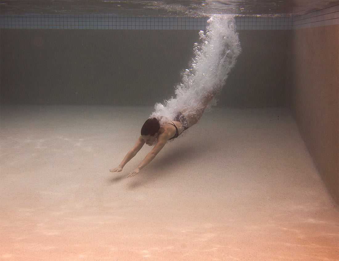 In Francesca Owen's "Self Reflection 01," a person is seen diving underwater in a swimming pool. The diver, clad in a swimsuit, has both arms extended forward, leaving a trail of bubbles as they enter the water. The scene mirrors the fine art paper quality and reveals the pool’s tiled wall in the background.