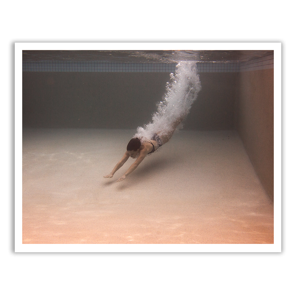 A person is diving underwater in a swimming pool, streamlined with arms extended forward. Bubbles trail behind them, highlighting the motion. The clear water reveals the pool's tiled wall in the background, creating an image reminiscent of Francesca Owen's hand-signed fine art piece "Self Reflection 01".