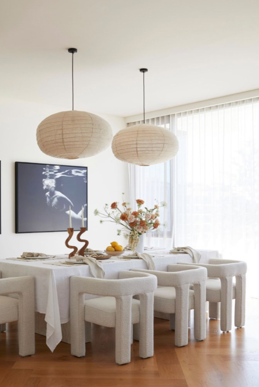 A modern dining room with light wood flooring features a large table covered with a white tablecloth, surrounded by eight cushioned chairs. Two large, round pendant lights hang above the table, which has a floral centerpiece and two candles. Wall art titled "Hold On" by Francesca Owen on fine art paper, large windows with vertical blinds, and natural light complete the space.