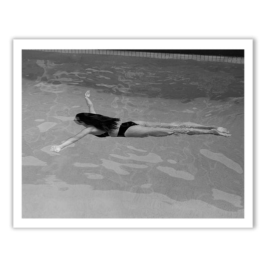 ‘Self Reflection 16’ by Francesca Owen captures a black and white image of a person floating face-up in a swimming pool, arms spread wide, with long hair flowing in the water. The surface of the pool reflects light while the tiled edge is visible at the top of the image. This fine art print is created using archival pigments to ensure lasting beauty.