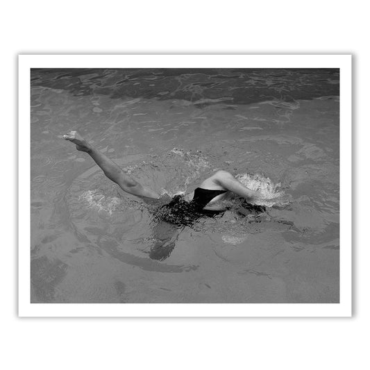 A black-and-white photograph titled "Self Reflection 18" by Francesca Owen captures a person swimming in a pool. One arm is extended out of the water, and one leg is kicking just above the surface. The person's head is submerged, surrounded by ripples. Printed with archival pigments on fine art paper, the overall composition emphasizes motion and fluidity.