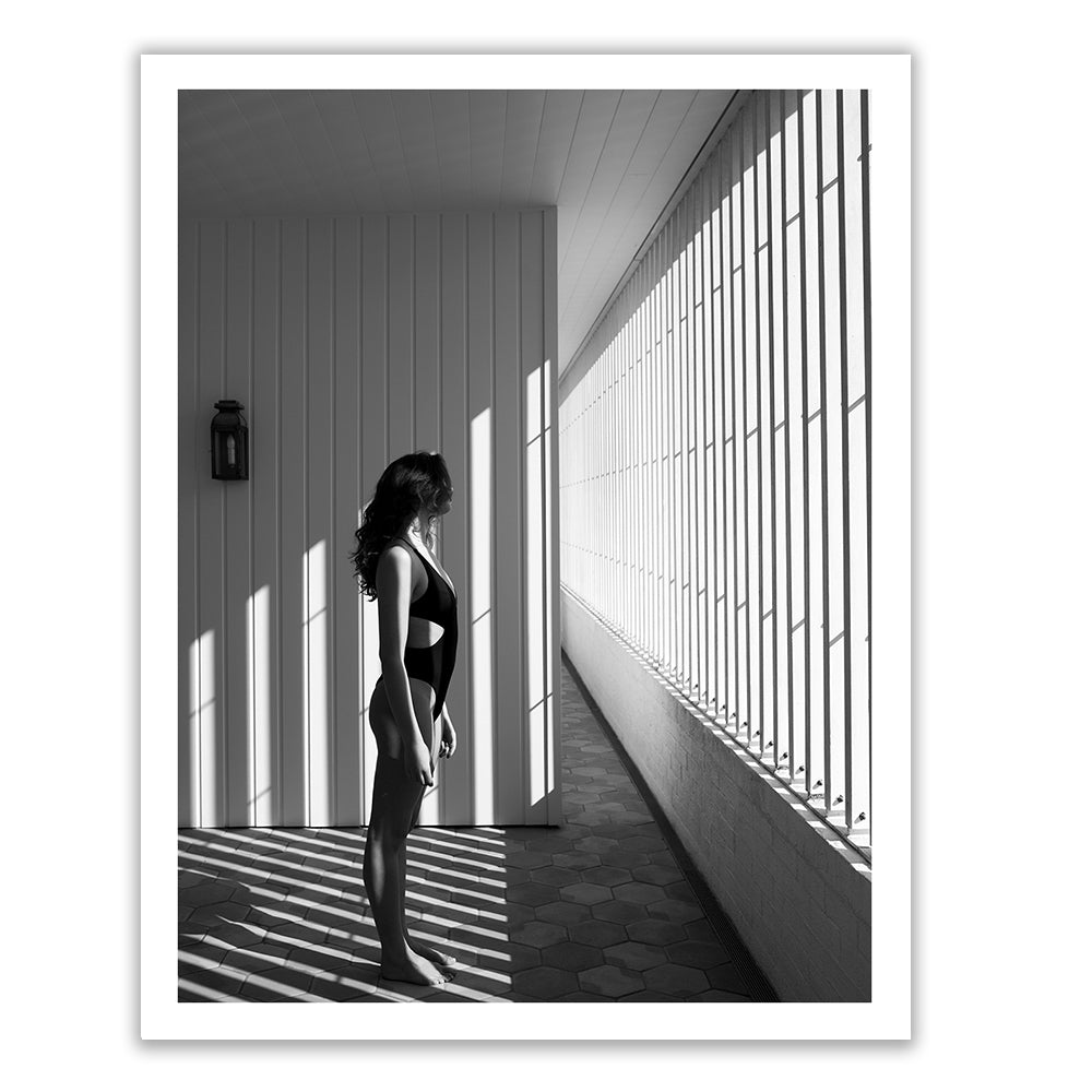 Self Reflection 10" by Francesca Owen features a striking black and white photo of a woman standing on a sunlit balcony, clad in a swimsuit. The woman faces a wall where the vertical shadows from the railing form an artistic pattern on both the floor and her body. This exquisite piece is printed on fine art paper with archival pigments and includes a certificate of authenticity.