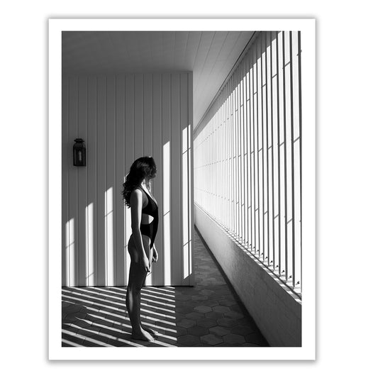 Self Reflection 10" by Francesca Owen features a striking black and white photo of a woman standing on a sunlit balcony, clad in a swimsuit. The woman faces a wall where the vertical shadows from the railing form an artistic pattern on both the floor and her body. This exquisite piece is printed on fine art paper with archival pigments and includes a certificate of authenticity.