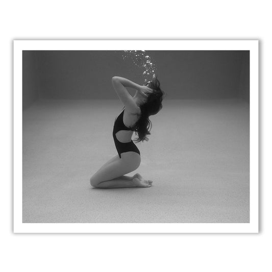 Self Reflection 11 by Francesca Owen features a captivating black-and-white underwater photo of a woman kneeling on her right knee, arching her back in a one-piece swimsuit. Bubbles rise above her head as she places her hands on her head, and her long hair flows gracefully through the water. Printed with archival pigments on fine art paper, this piece ensures lasting beauty.