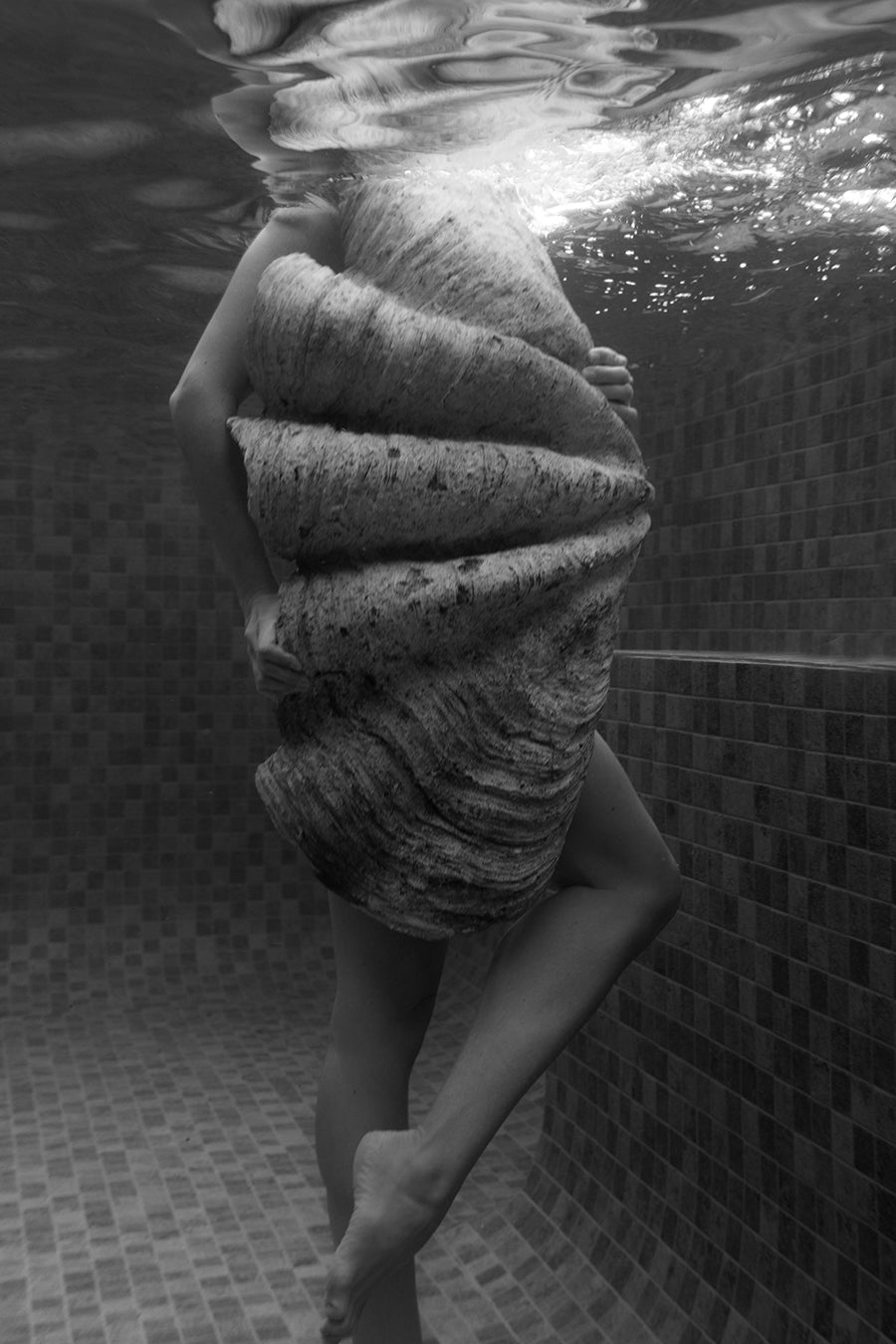 In Francesca Owen’s "Shell & She #2," a person stands in a tiled pool, holding a large shell before their torso. The black and white photo captures their face above water, with bent knee and submerged legs visible.