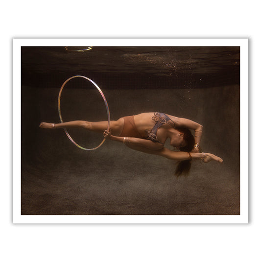In "Show Time Part 3, #1" by Francesca Owen, a performer elegantly maneuvers a hula hoop underwater, showcasing smooth movement and perfect balance. The water imparts a tranquil, ethereal atmosphere to the scene, similar to fine art prints created with archival pigments.