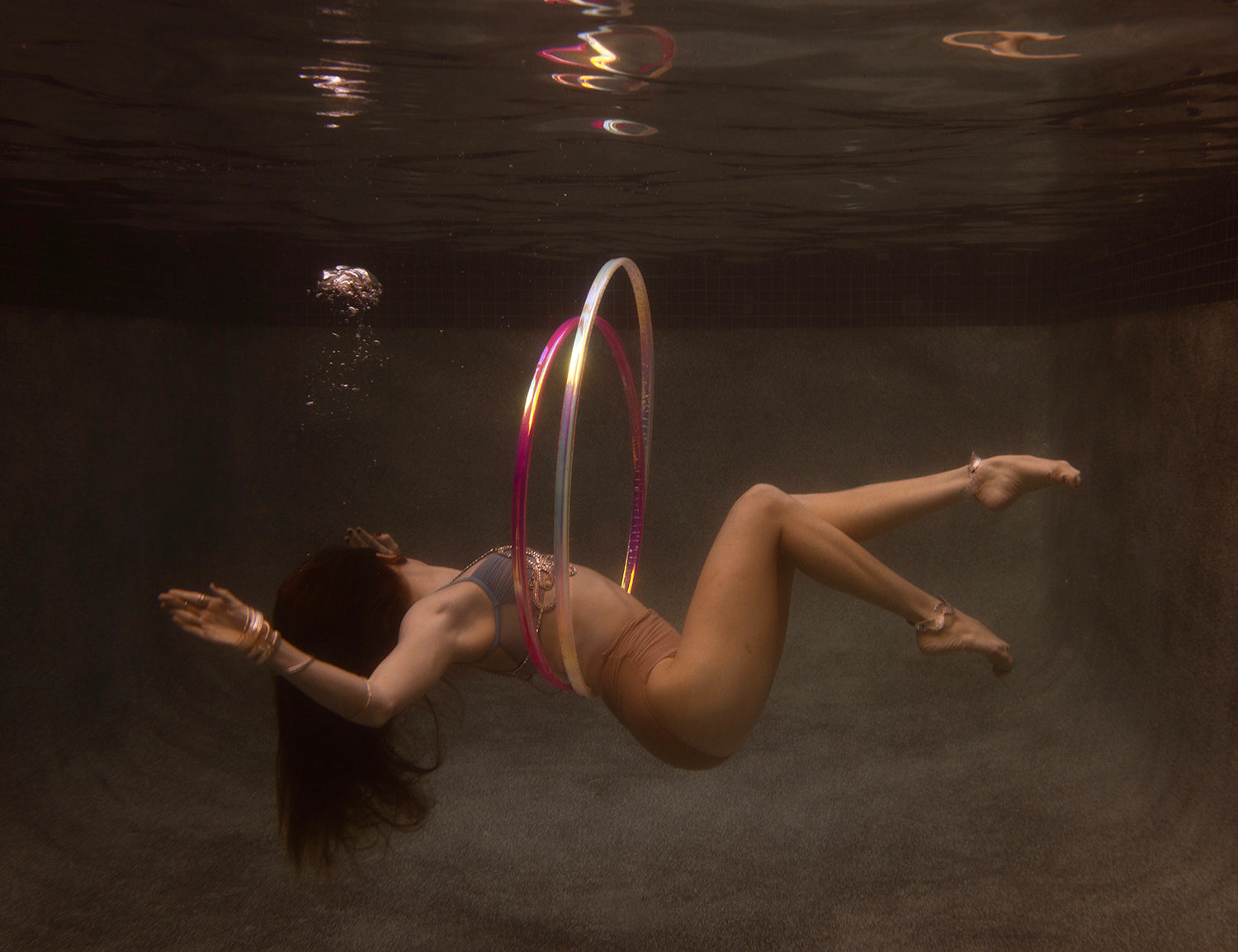 A woman floats underwater, her back elegantly arched and arms outstretched. She wears a bikini amidst a vibrant display of colorful hula hoops. Captured on fine art paper using archival pigments, bubbles ascend above her in the dim water, crafting an ethereal scene. This artwork is titled "Show Time Part 3, #2" by Francesca Owen.