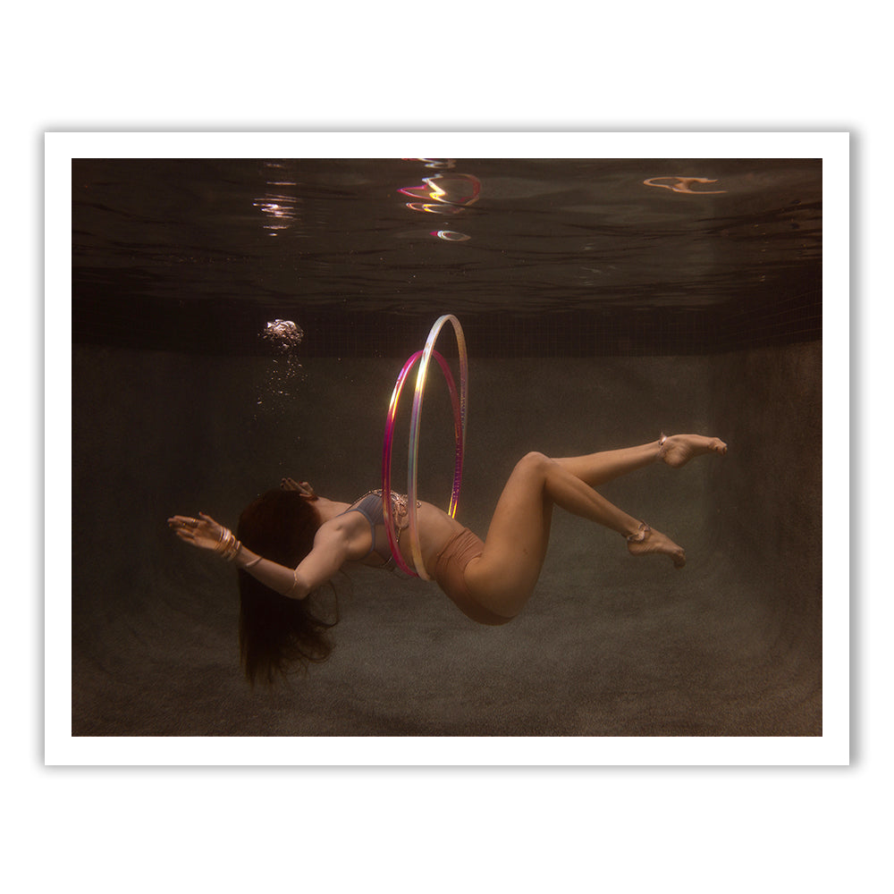Gracefully hula hooping with a colorful hoop, a woman with long hair swims underwater. Bubbles rise around her in the pool, adding a dynamic element to the serene setting as if captured on fine art paper using archival pigments in "Show Time Part 3, #2" by Francesca Owen.