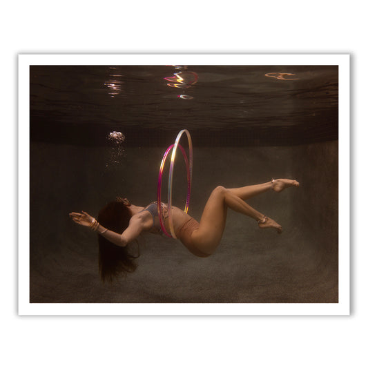Gracefully hula hooping with a colorful hoop, a woman with long hair swims underwater. Bubbles rise around her in the pool, adding a dynamic element to the serene setting as if captured on fine art paper using archival pigments in "Show Time Part 3, #2" by Francesca Owen.