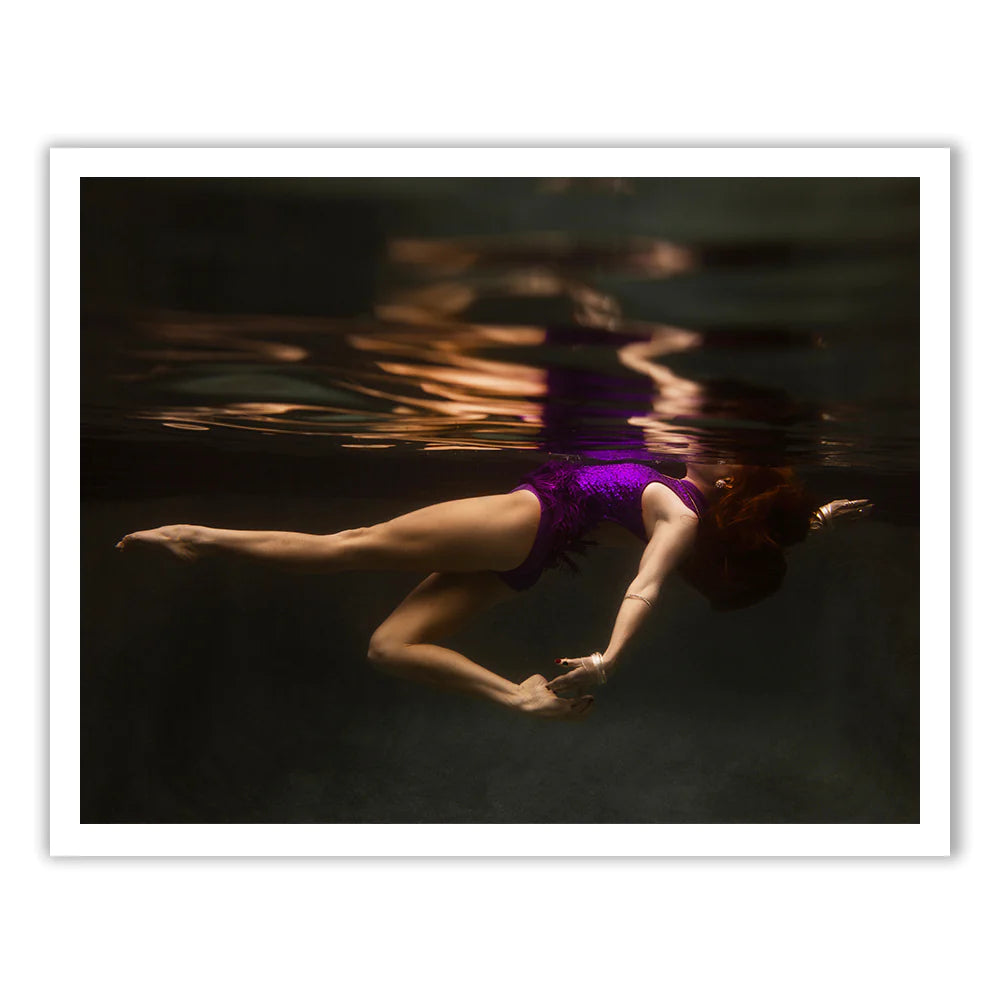 A woman wearing the Show Time Part 2, #2 swimsuit by Francesca Owen gracefully floats underwater, her arms extended and hair gently flowing. The low light creates soft reflections on the water's surface, adding a serene and artistic ambiance reminiscent of fine art paper prints crafted with archival pigments.